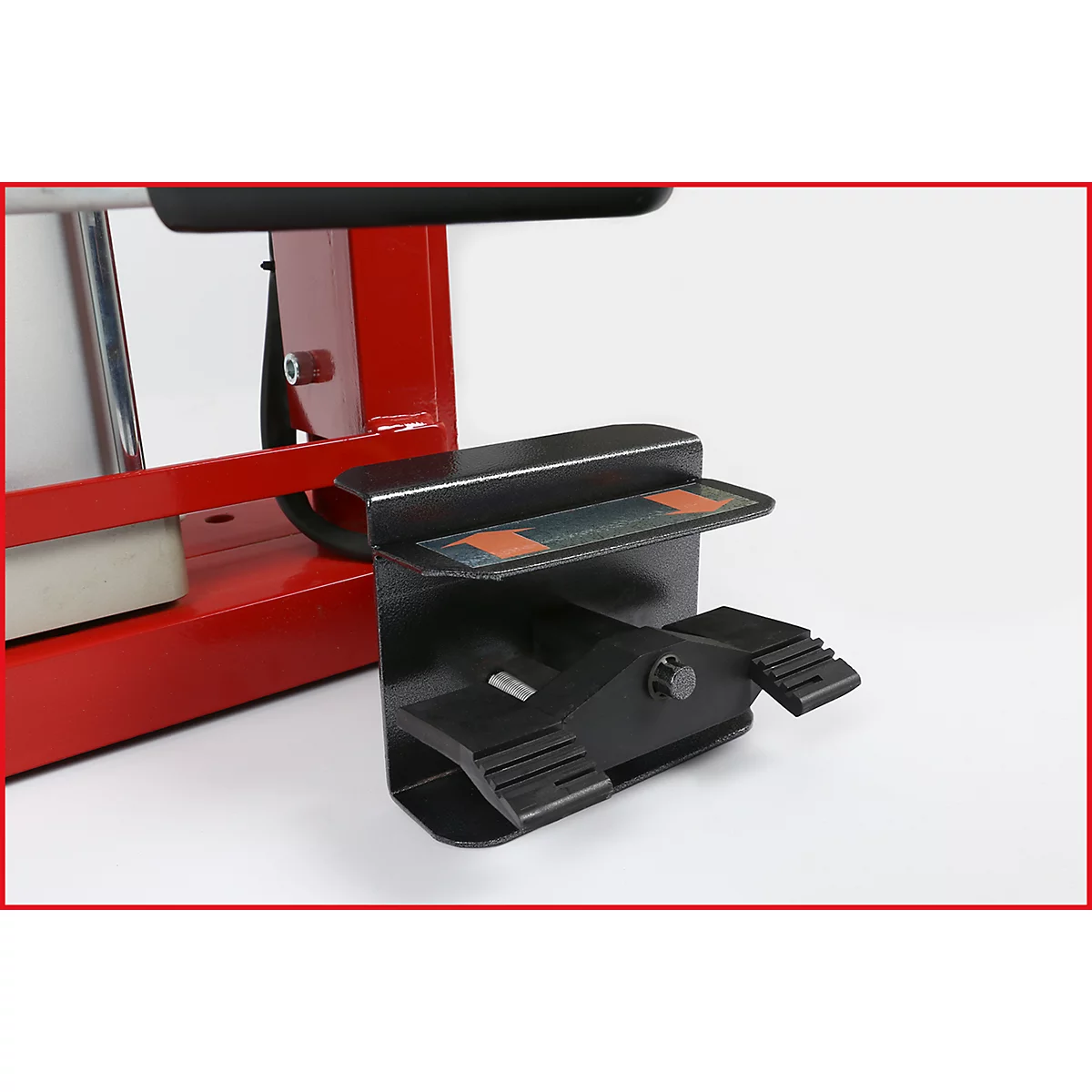 Detailaufnahme eines schwarzen Fußpedals mit einer rot-schwarzen Maschine im Hintergrund. Das Pedal ist an einer Halterung befestigt.